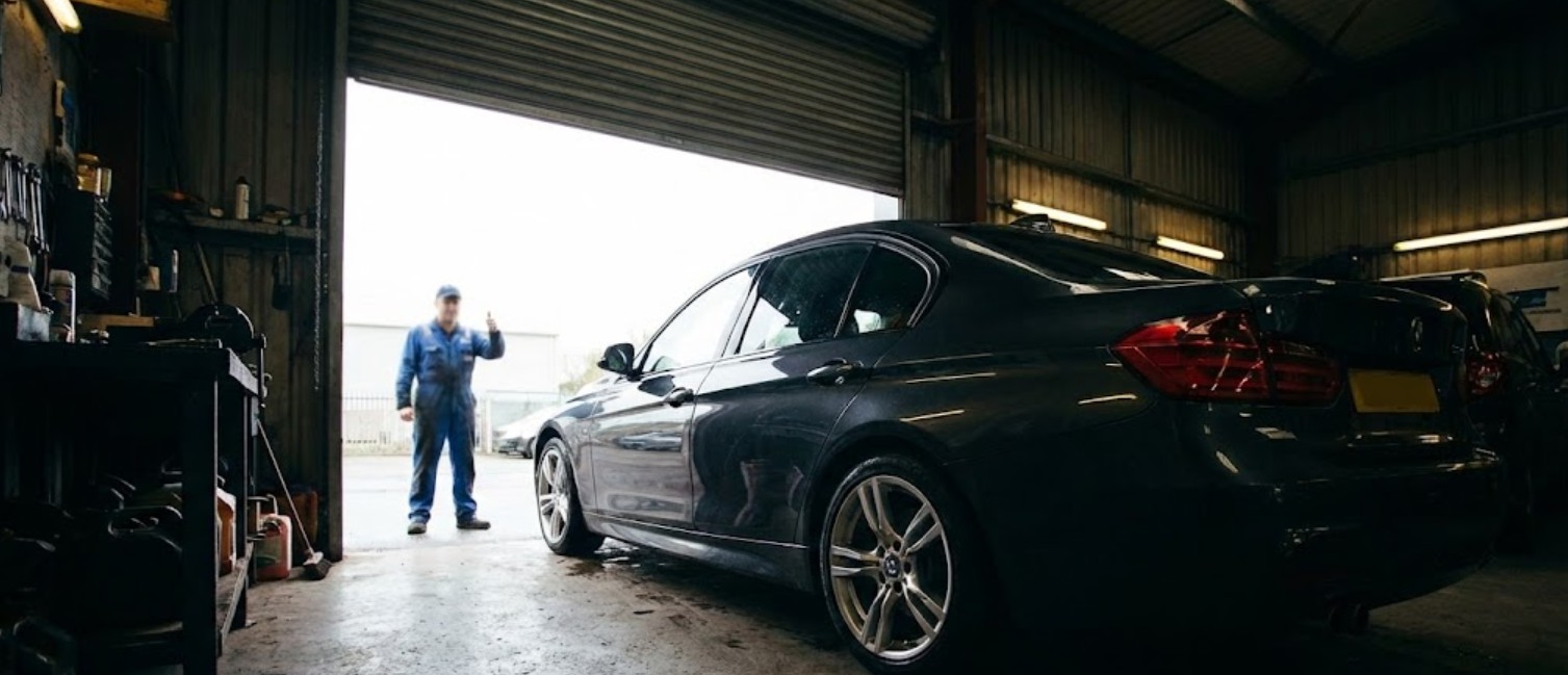 Black car parked inside a dimly lit garage with a mechanic in blue overalls outside giving a thumbs-up.