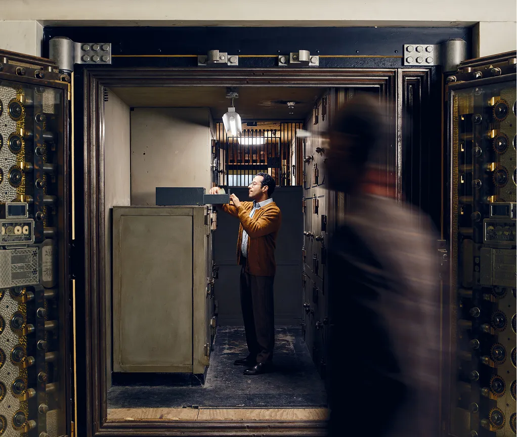 Man in brown jacket standing inside a large bank vault, opening a safe deposit box, with a blurred figure walking past the open vault door.