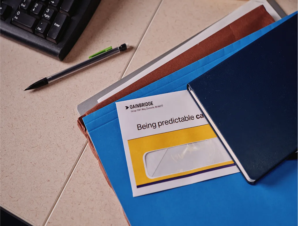 Desk scene with a black pen, keyboard, and folder holding an envelope from Gainbridge partially covered by a dark blue notebook.