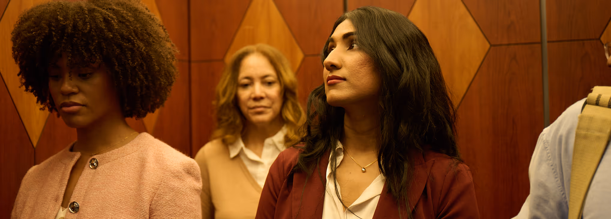 Four people standing inside an elevator with wood-paneled walls, with three fully visible and one partially seen.
