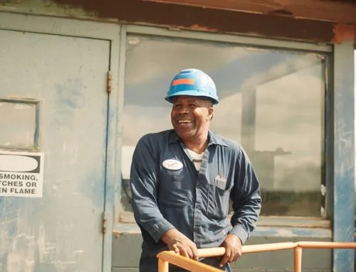 Smiling male electrician wearing a blue helmet and navy work shirt leaning on a railing outside an industrial building.