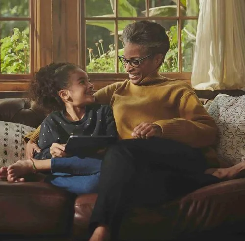 Smiling older woman in glasses and mustard sweater sitting on sofa with young girl holding a tablet, both looking at each other warmly.