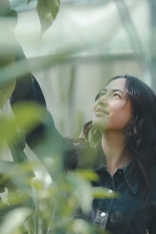 Woman with long dark hair wearing a denim jacket reaching up toward green leaves outdoors.