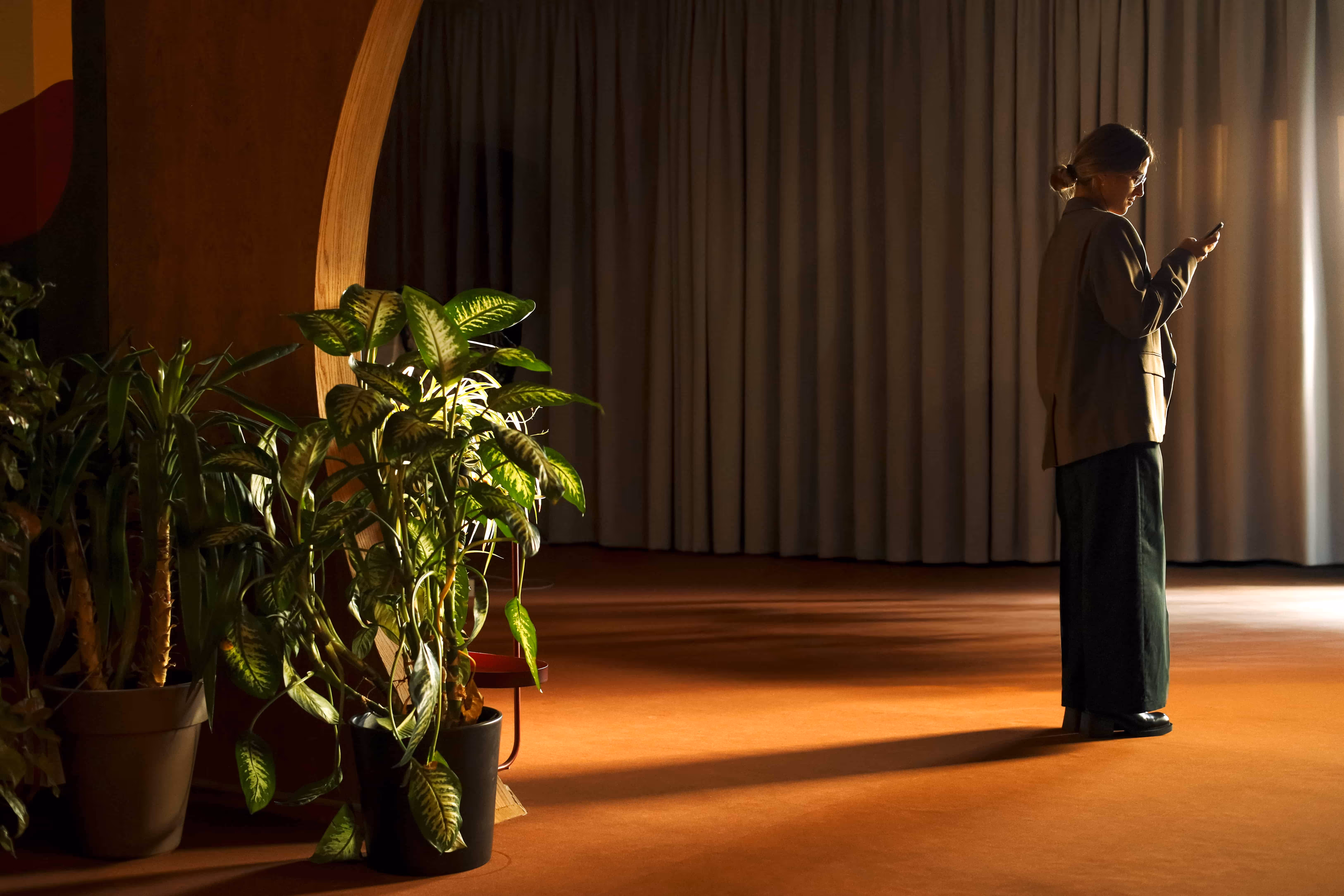 Woman standing in a dimly lit room, looking at her phone with large potted plants beside her and heavy curtains in the background.