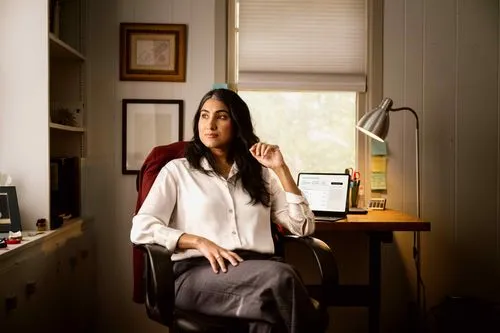 Woman in a white blouse and gray skirt sitting in an office chair, looking out a window with a desk, laptop, and lamp behind her.
