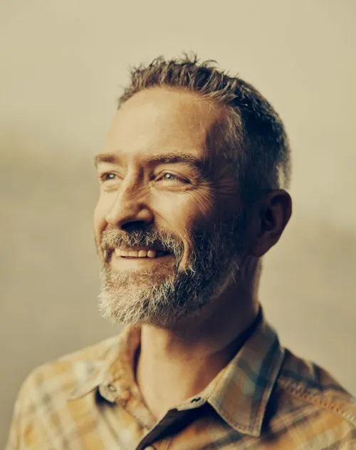 Smiling middle-aged man with gray hair and beard wearing a plaid shirt looking to the side.