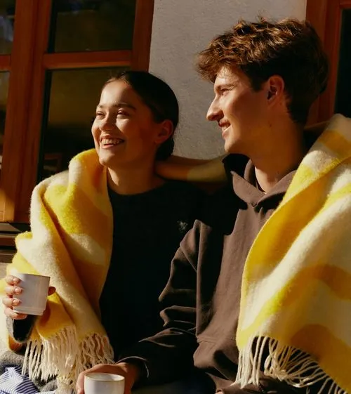 Young couple wrapped in a yellow and white striped blanket, sitting close and smiling while holding white cups.