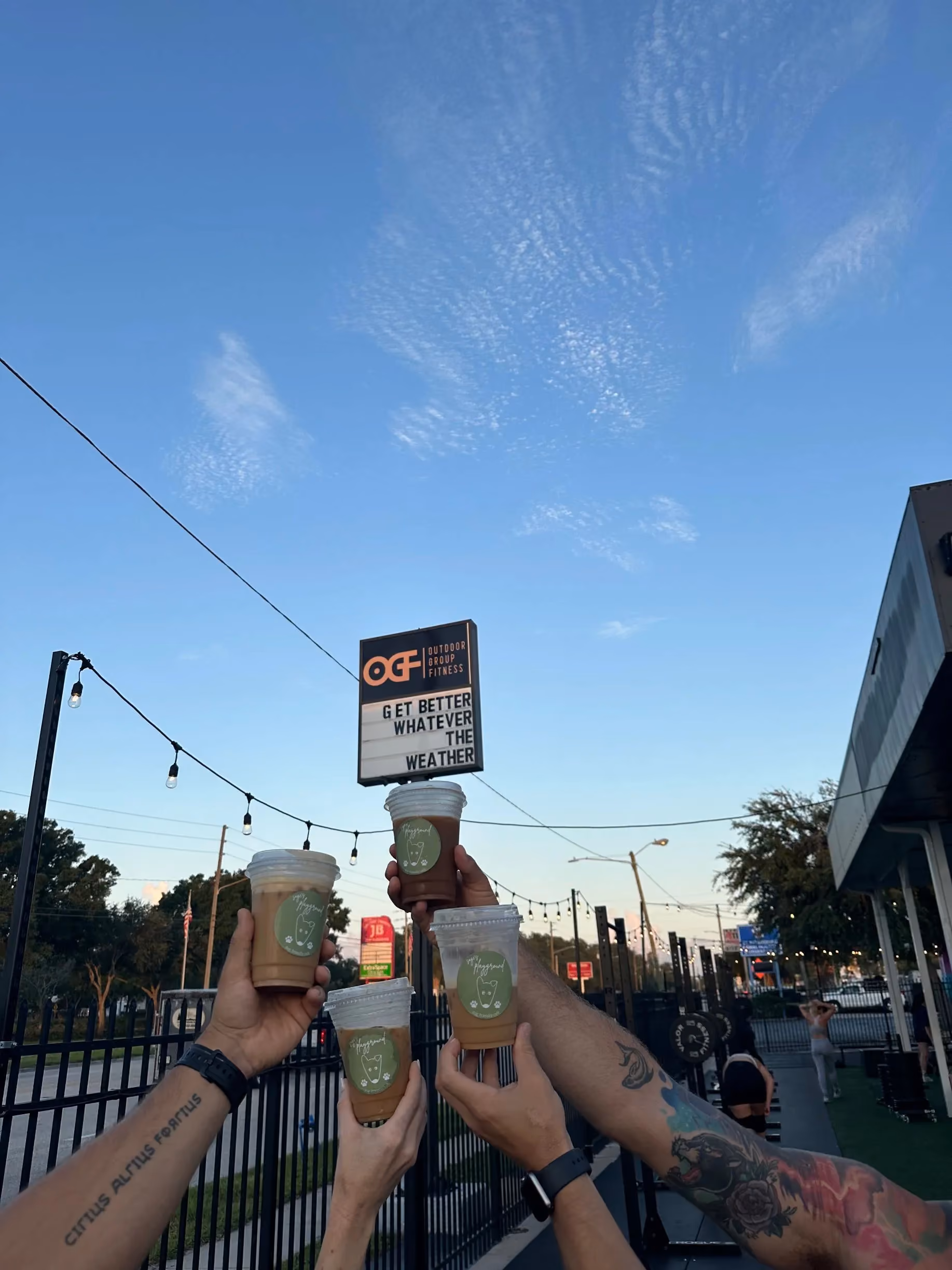 Three guests holding Sage's Playground coffee in front of the sign at an Outdoor Group Fitness event in Tampa Bay
