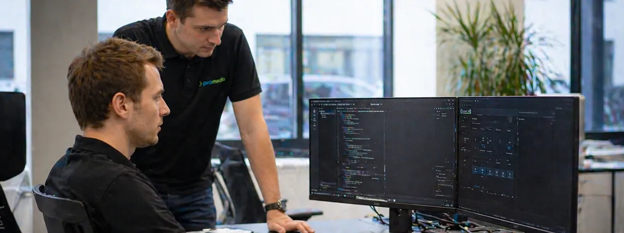 Zwei Promedia-Techniker planen die AV-Systemintegration für einen Konferenzraum an einem Dual-Monitor-Arbeitsplatz in Hockenheim.