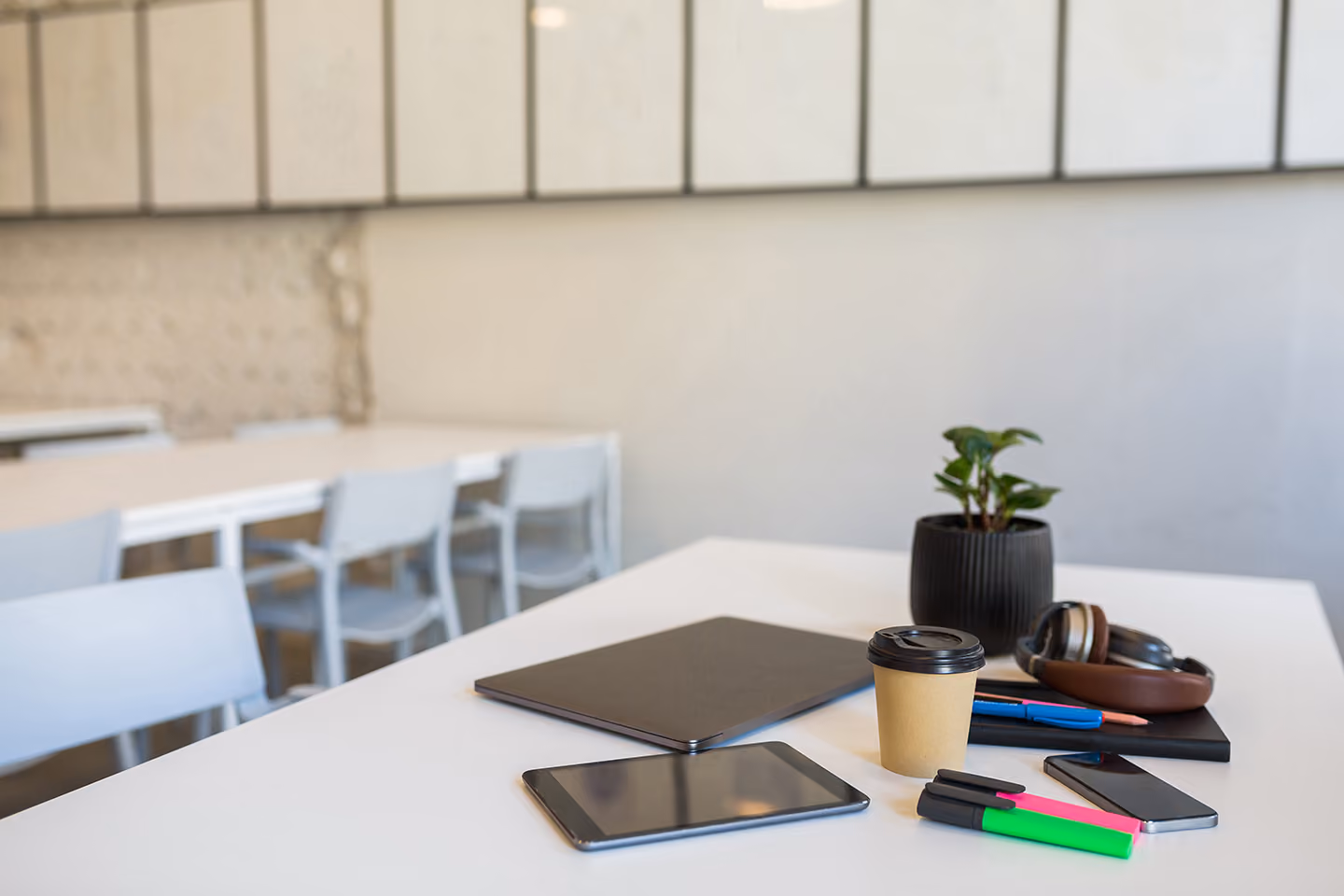 White office table with tablet, closed laptop, coffee cup, headphones, pens, highlighters, smartphone, notebook, and a small potted plant in a modern workspace.