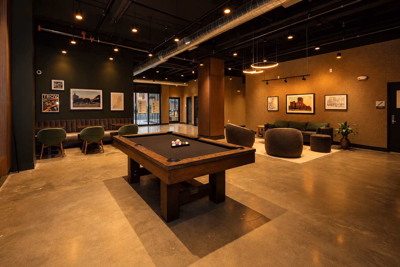 Modern recreation room with a black felt pool table in the center, three green chairs and framed pictures on the dark wall to the left, and a cozy seating area with round gray chairs, a couch, and framed artwork on the beige wall to the right.
