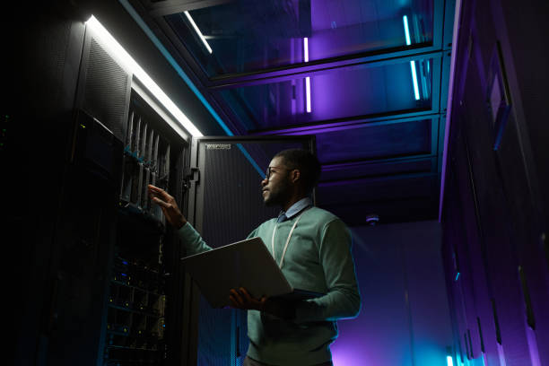 Man holding a laptop while inspecting a server rack in a dimly lit data center with purple and blue lighting.