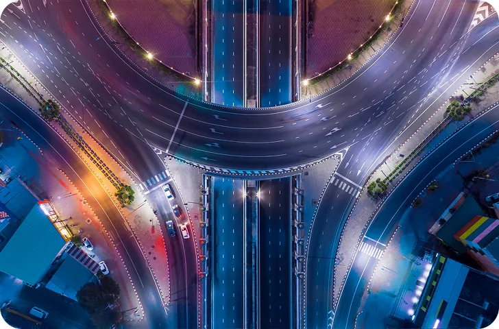 Aerial view of a wide, illuminated city intersection with multiple lanes and few cars driving at night.