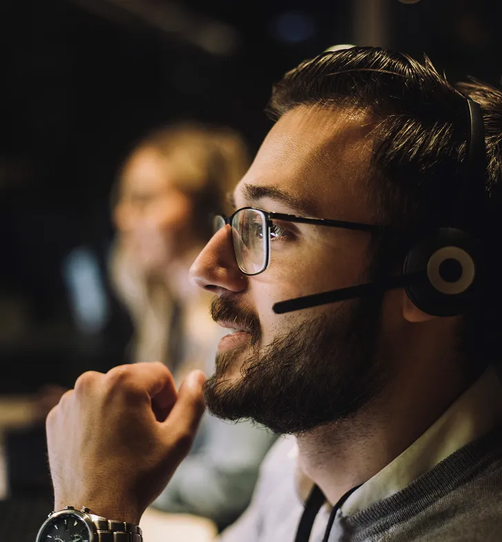 Man with glasses and headset smiling thoughtfully in a dimly lit office environment.