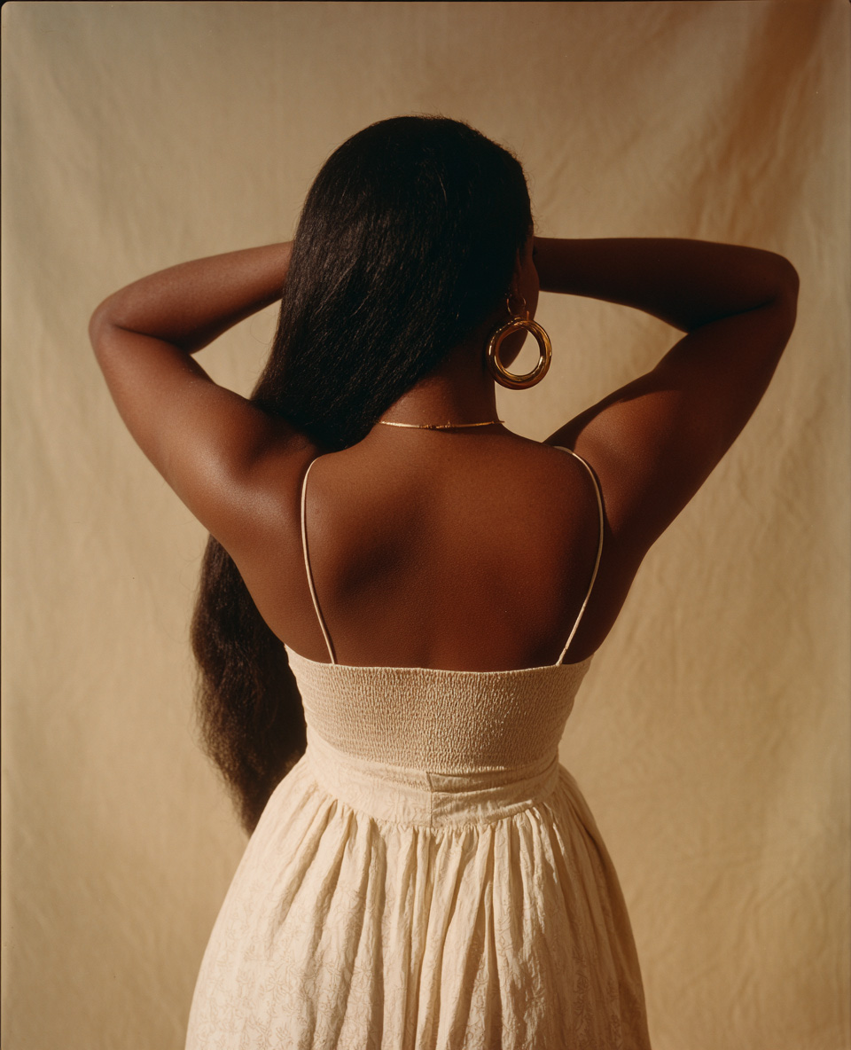 Rückenansicht einer Frau mit langen dunklen Haaren, die eine cremefarbene Sommerkleidung und große goldene Ohrringe trägt, Arme hinter dem Kopf verschränkt.