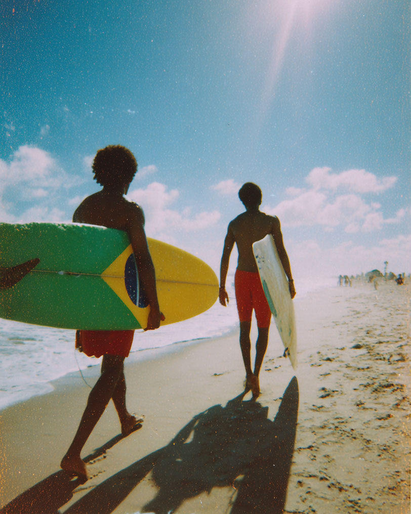 Zwei Surfer mit Surfboards gehen am sonnigen Strand entlang, einer mit einem Board in den Farben der brasilianischen Flagge.