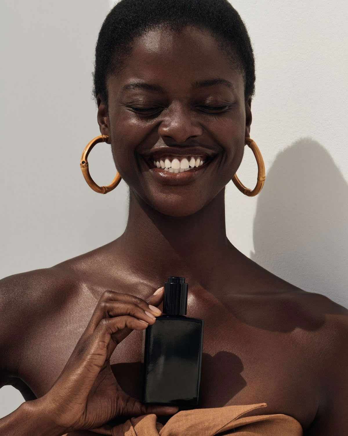 Lächende Frau mit großen Bambus-Ohrringen hält eine schwarze Kosmetikflasche vor ihrer Brust.