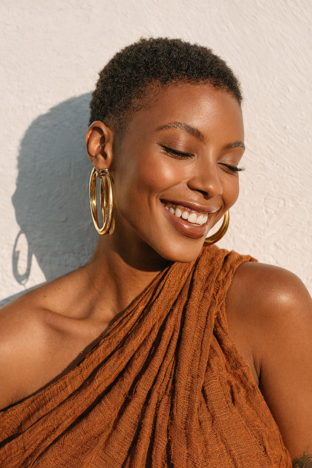 Porträt einer lächelnden Frau mit kurzen lockigen Haaren, großen goldenen Creolen und einem orangefarbenen einseitigen Oberteil vor einer hellen Wand.