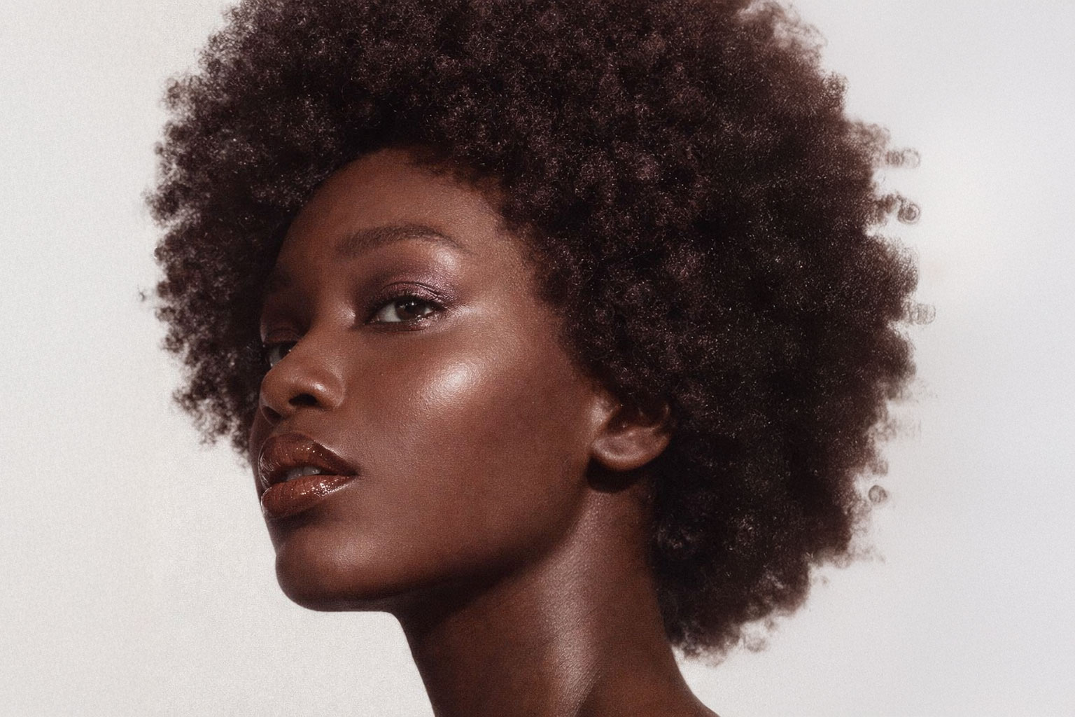 Nahaufnahme einer Frau mit natürlichem Afro-Haar und schimmerndem Lippenstift vor hellem Hintergrund.