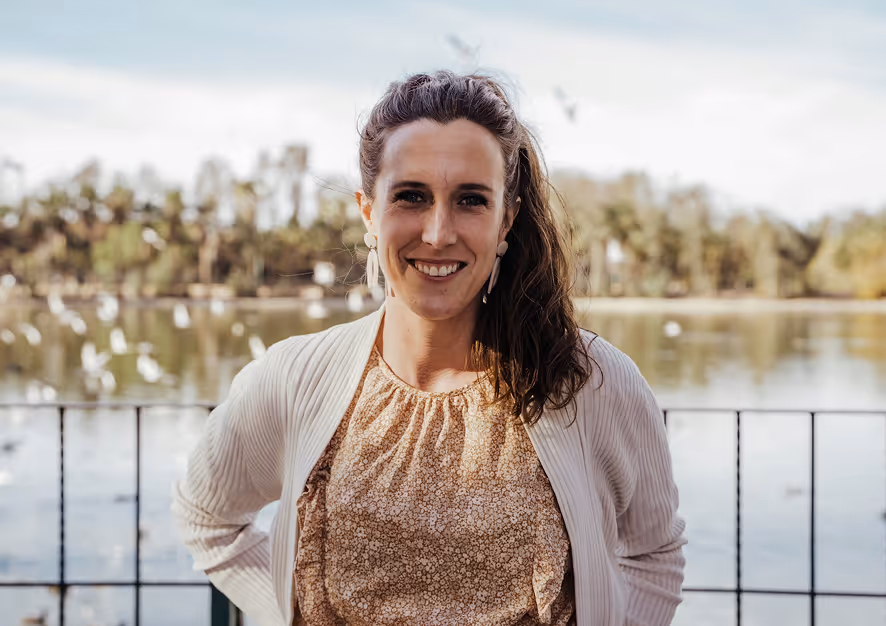 Smiling woman with long wavy hair standing outdoors by a lake with trees and birds in the background.