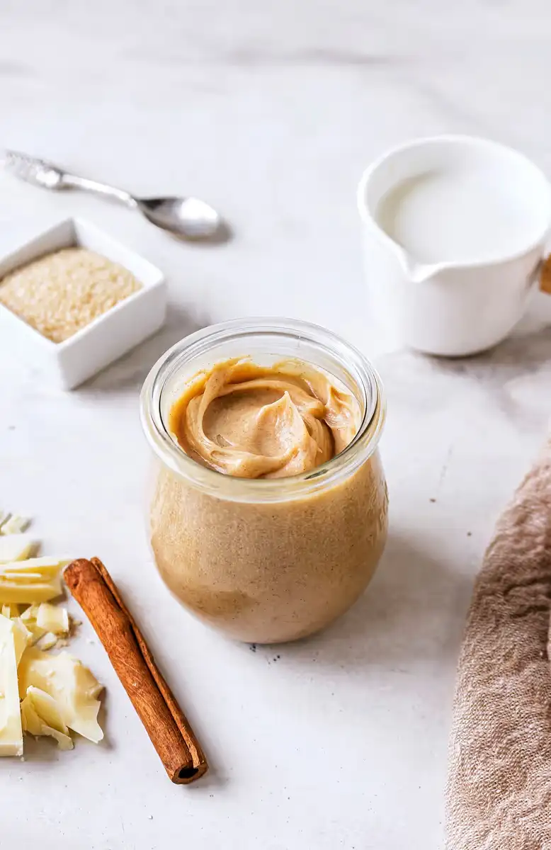 Jar of creamy almond butter surrounded by cinnamon stick, white chocolate shavings, raw sugar in a small dish, and a white creamer on a marble surface.