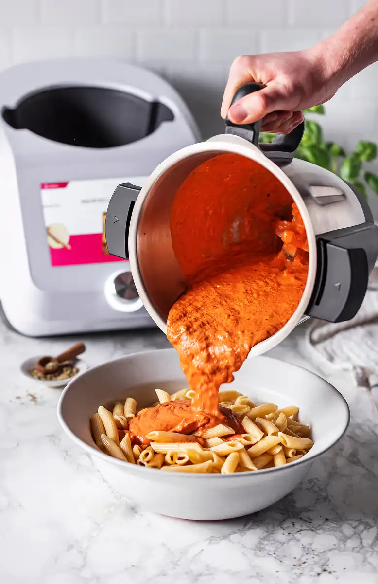 Hand pouring creamy red sauce from a metal container into a bowl of cooked penne pasta on a marble countertop.