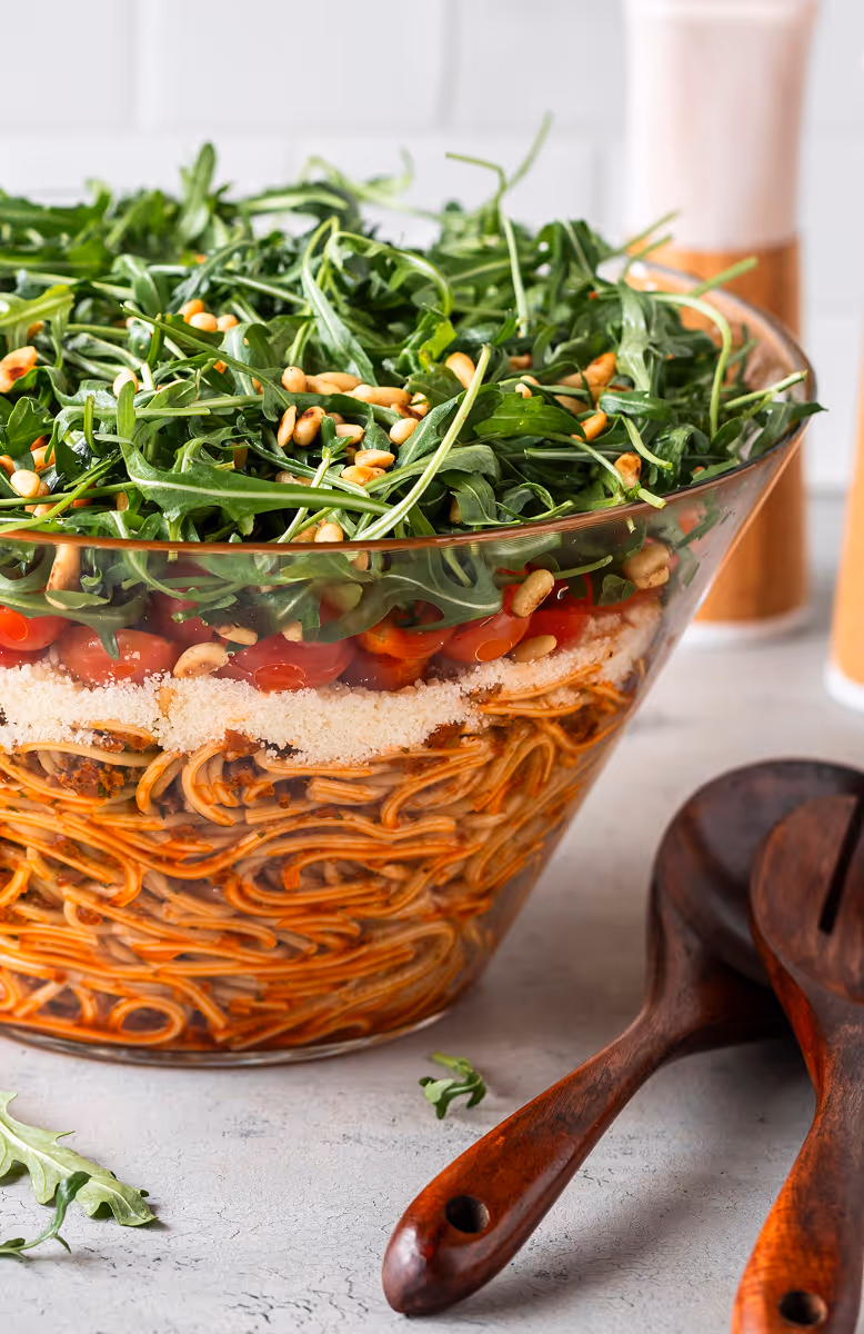 Layered spaghetti salad in a glass bowl topped with arugula and pine nuts, with wooden salad utensils beside it.