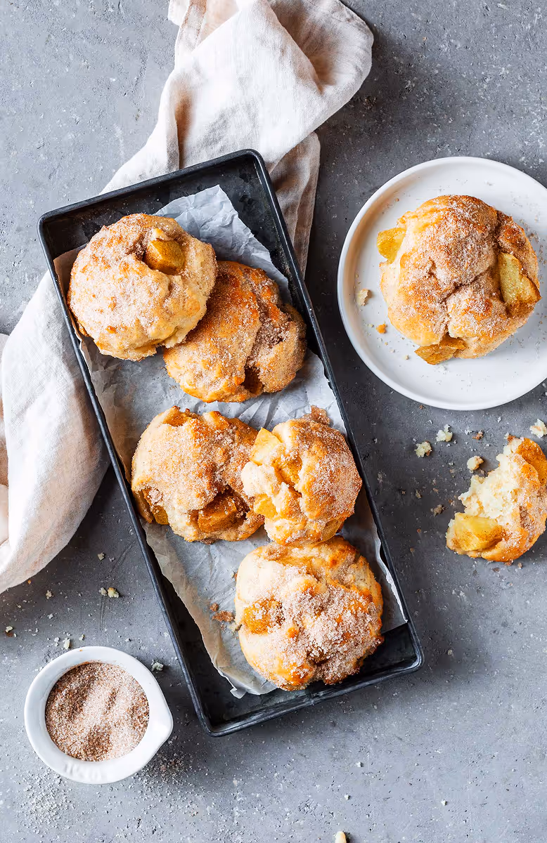 Five cinnamon sugar-coated baked apple fritters on parchment in a black tray next to one on a white plate and crumbs on a gray surface.