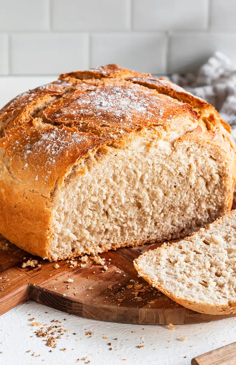 Freshly baked round loaf of bread with a slice cut, resting on a wooden cutting board with crumbs scattered around.