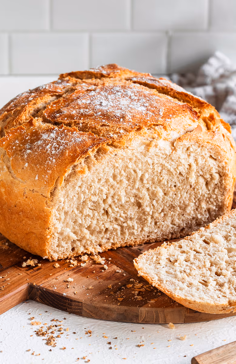 Freshly baked round loaf of bread with a slice cut, resting on a wooden cutting board with crumbs scattered around.