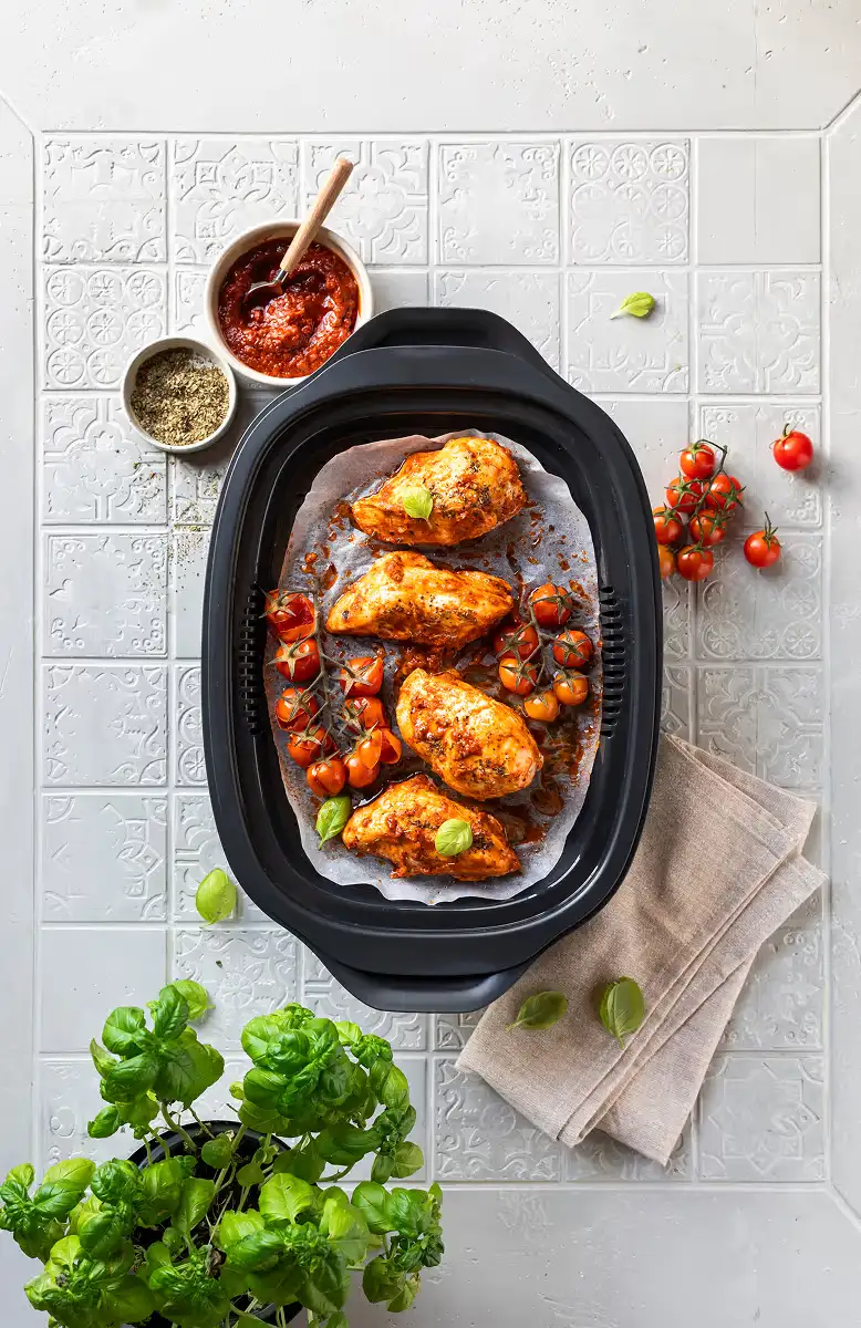 Cooked chicken breasts with cherry tomatoes and basil leaves in a black cooking tray on a tiled surface with herbs and tomato sauce beside it.