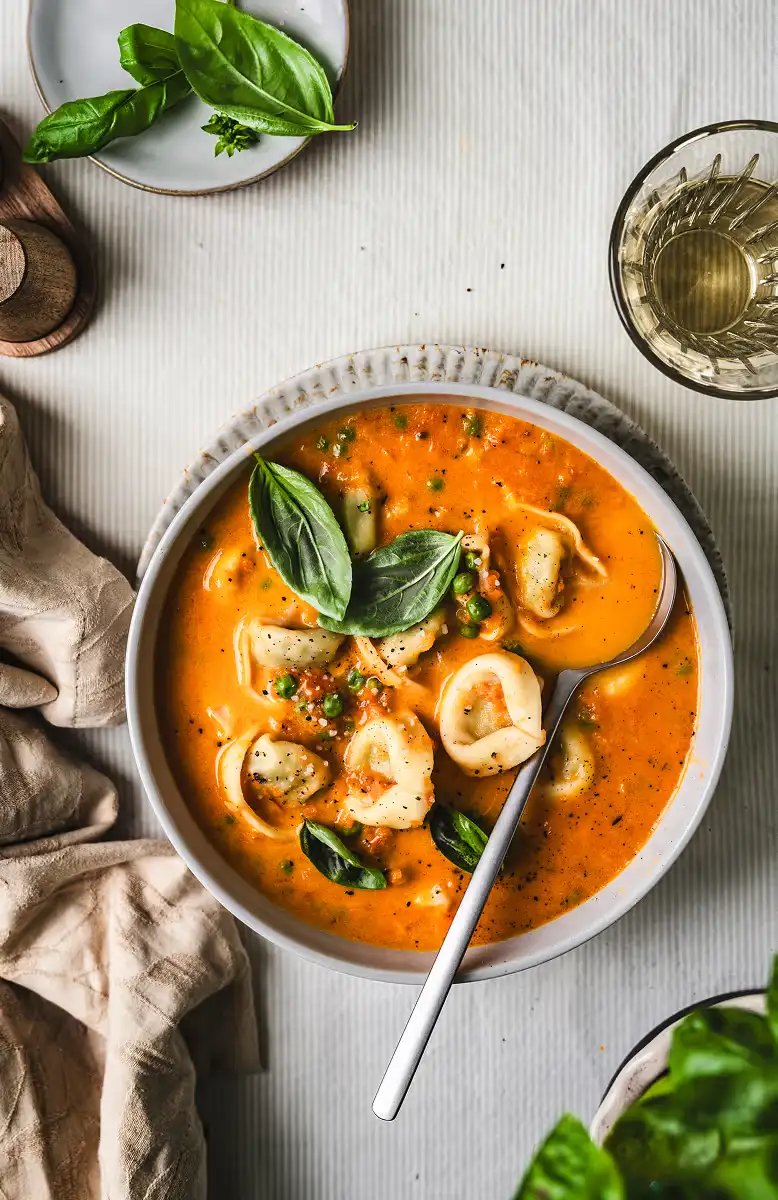Bowl of tomato-based tortellini soup garnished with fresh basil leaves and black pepper, with a spoon inside the bowl.