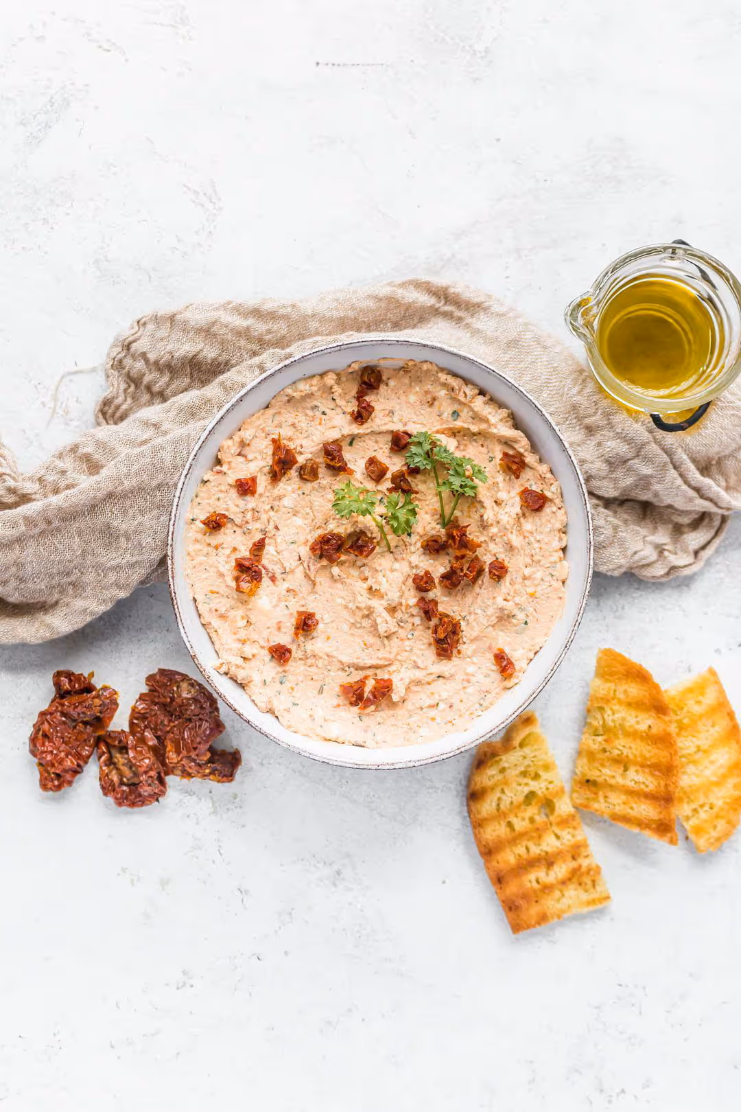 Creamy dip garnished with sun-dried tomatoes and parsley in a bowl, accompanied by grilled bread slices, sun-dried tomatoes, and a small glass jug of olive oil.