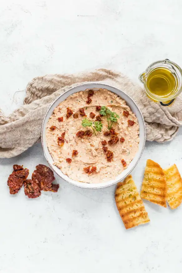 Creamy dip garnished with sun-dried tomatoes and parsley in a bowl, accompanied by grilled bread slices, sun-dried tomatoes, and a small glass jug of olive oil.