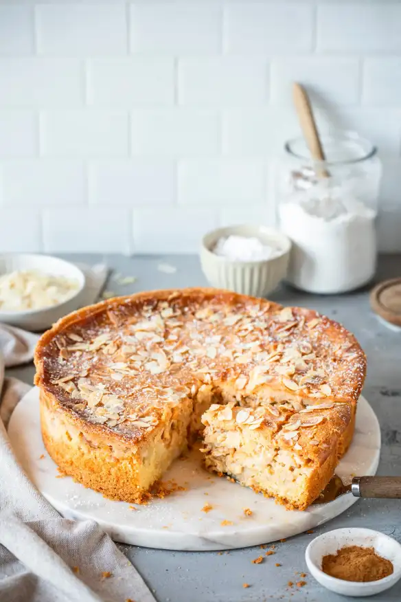 Slice of almond-topped apple cake on a white marble serving board with a cake server and baking ingredients in the background.