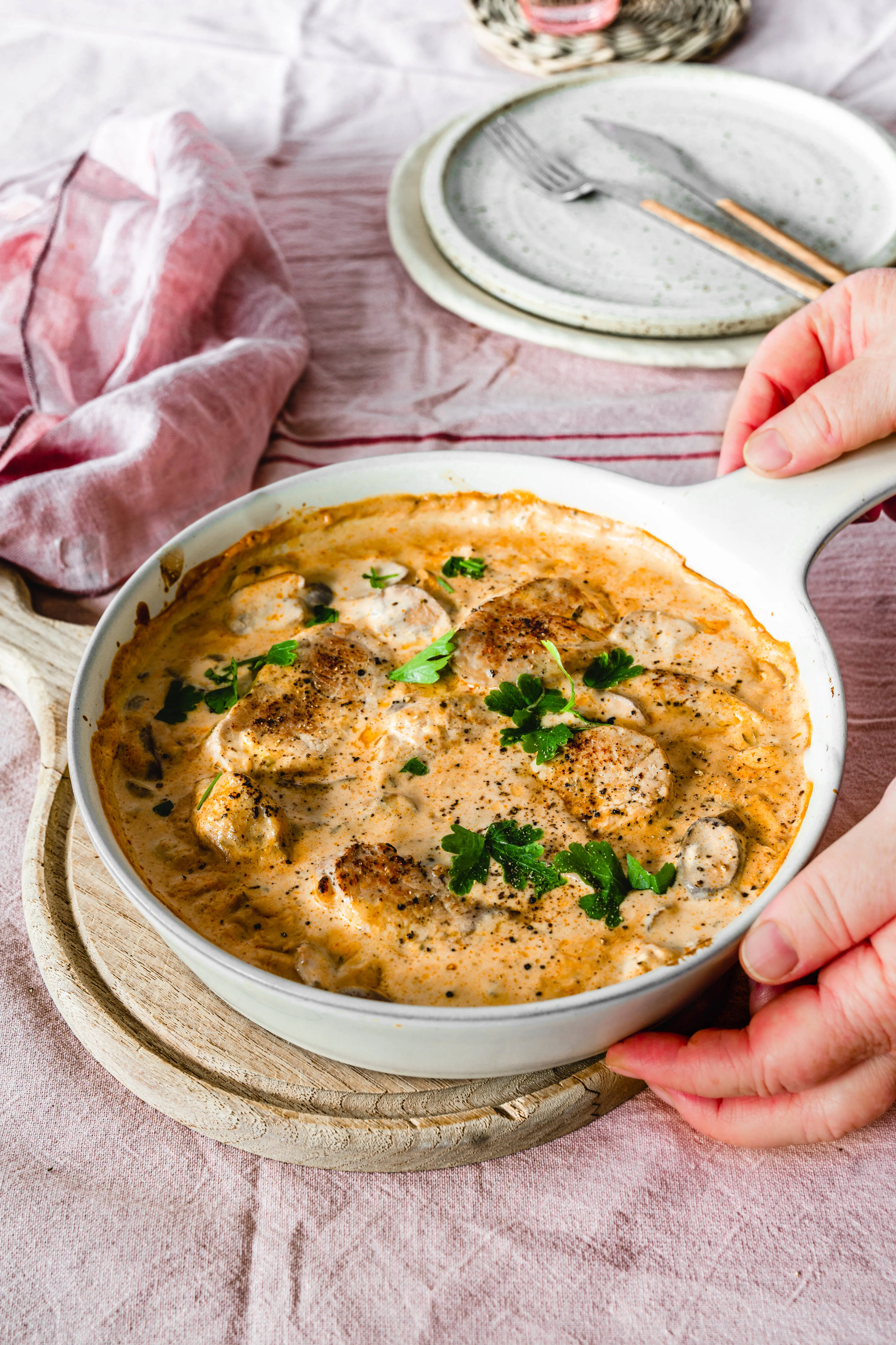 White pan with creamy pork medallions in mushroom sauce garnished with fresh parsley on a wooden trivet.