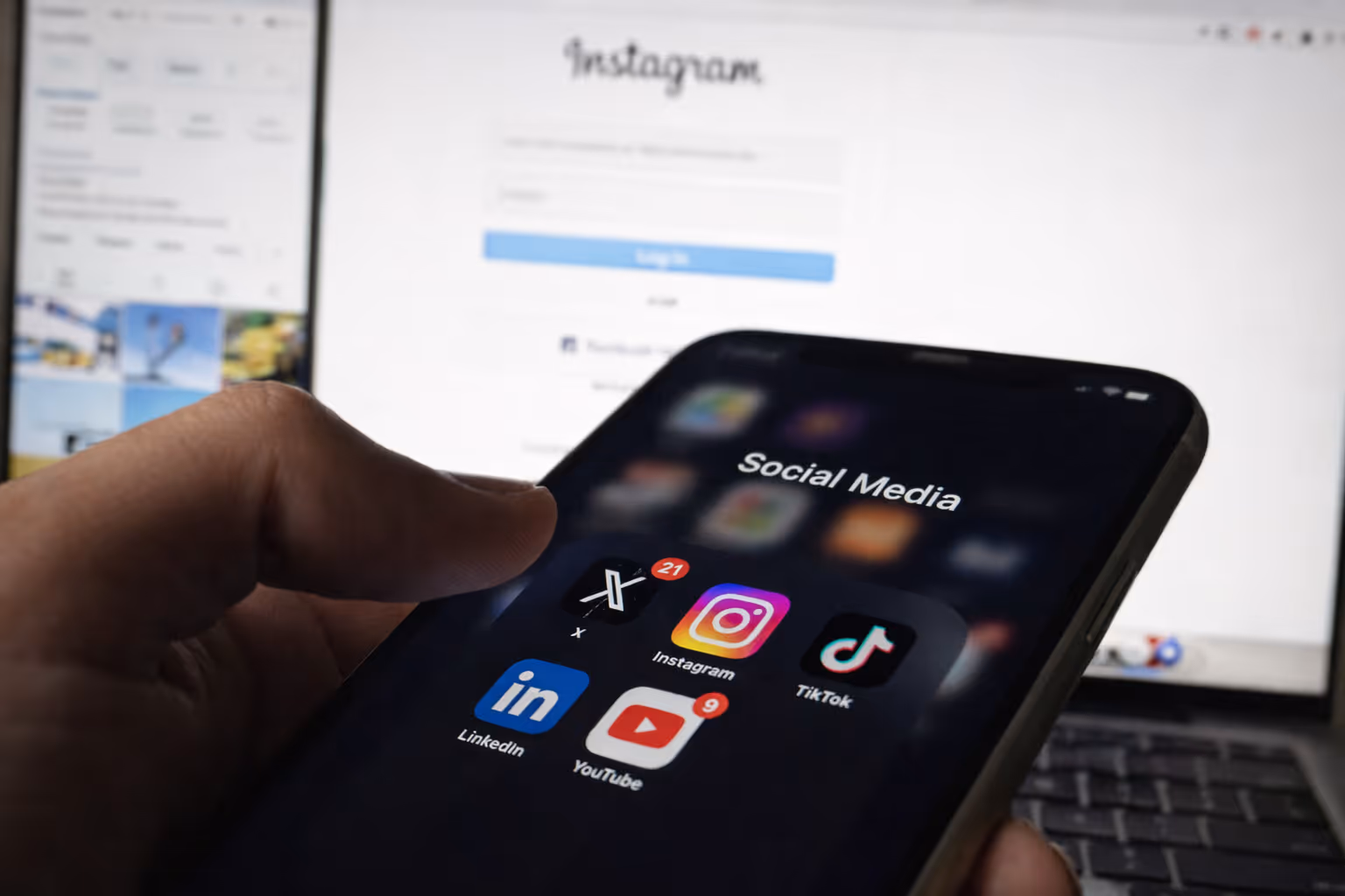 A close-up shot focuses on a person's thumb interacting with a smartphone screen displaying a folder titled "Social Media." The background shows a computer monitor with a blurred social media interface.