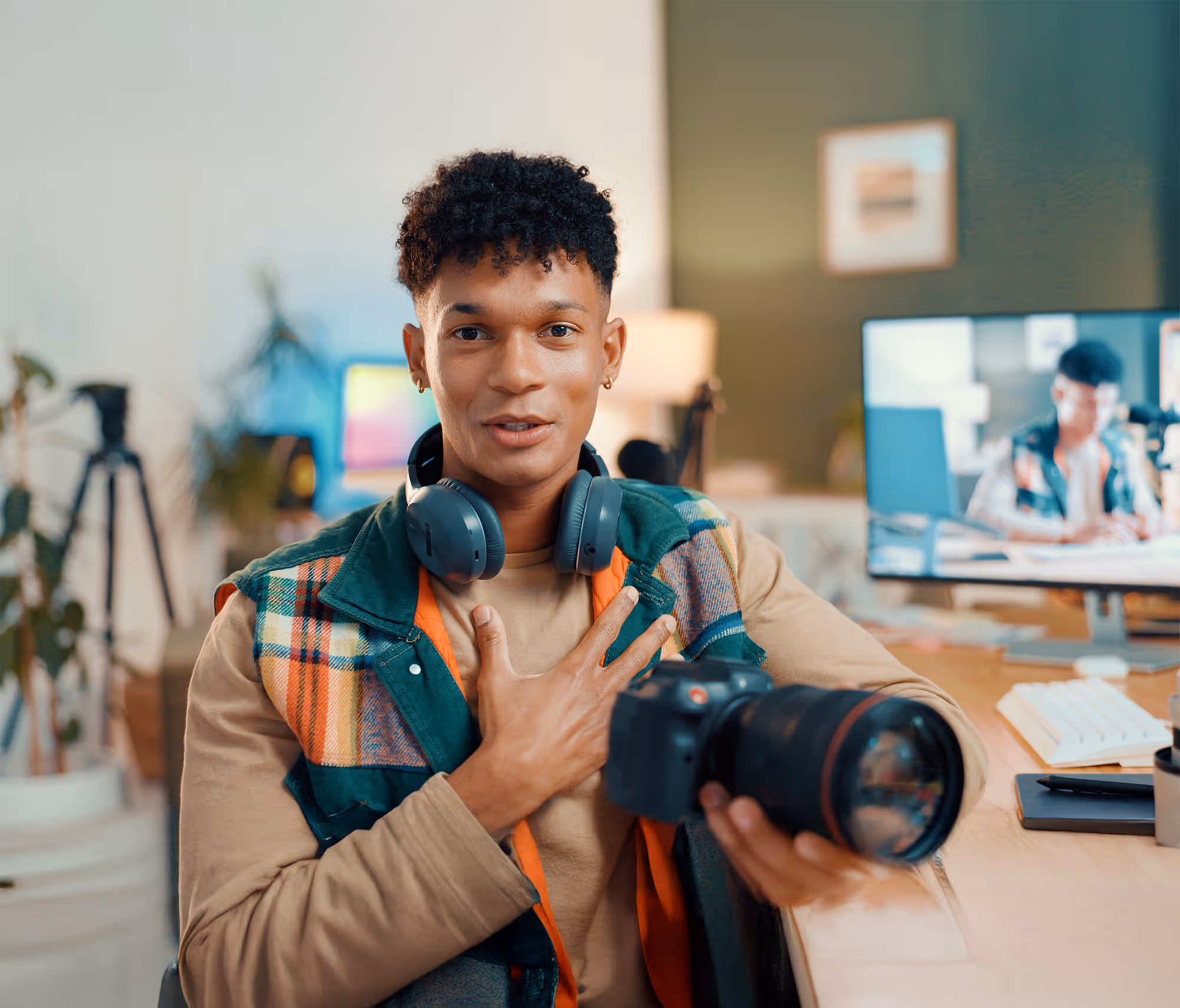 A young man with short, curly hair and a warm expression sits at a bright desk, presenting a professional DSLR camera to the viewer. He appears to be a content creator or photographer in a well-equipped home studio.