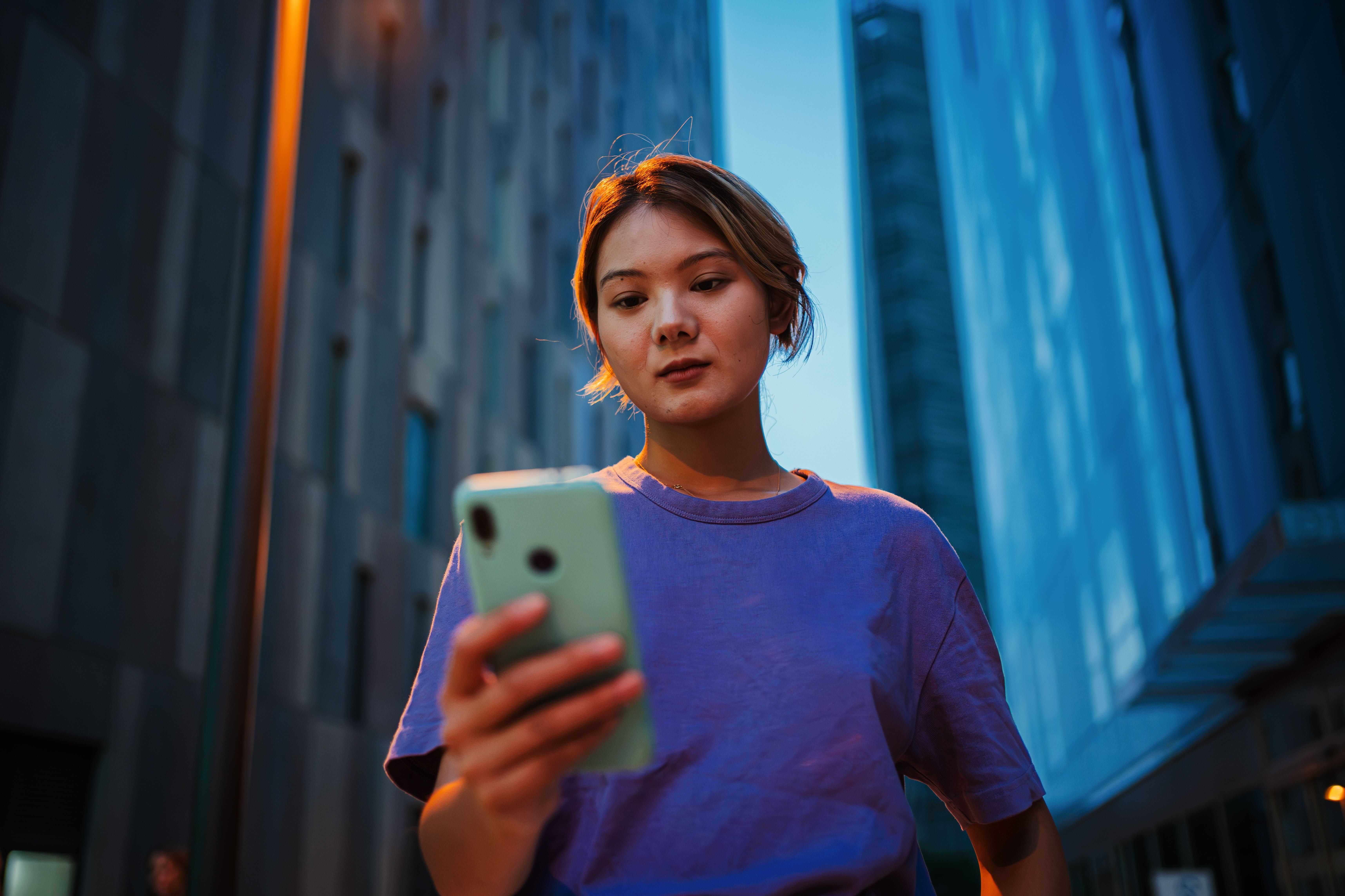  Gemini said A young woman with short hair stands in a modern city at dusk, her face illuminated by the glow of the smartphone she is holding. She is framed by towering glass skyscrapers that reflect the deep blue of the twilight sky.