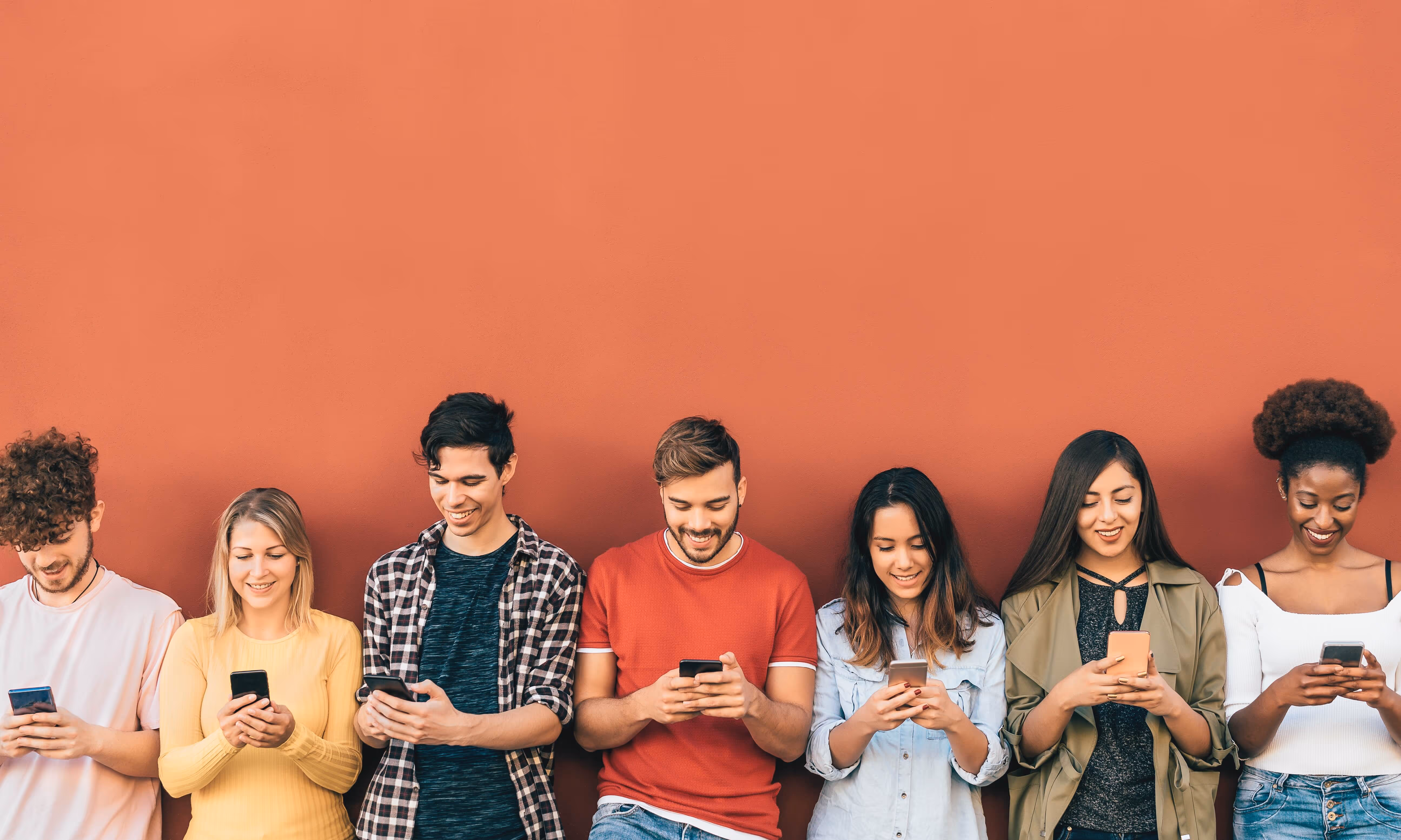 A group of seven diverse young adults stands in a horizontal line against a solid, reddish-orange wall, all looking down at their smartphones with smiling or pleasant expressions. The image is a wide shot with a large amount of empty space above the group.