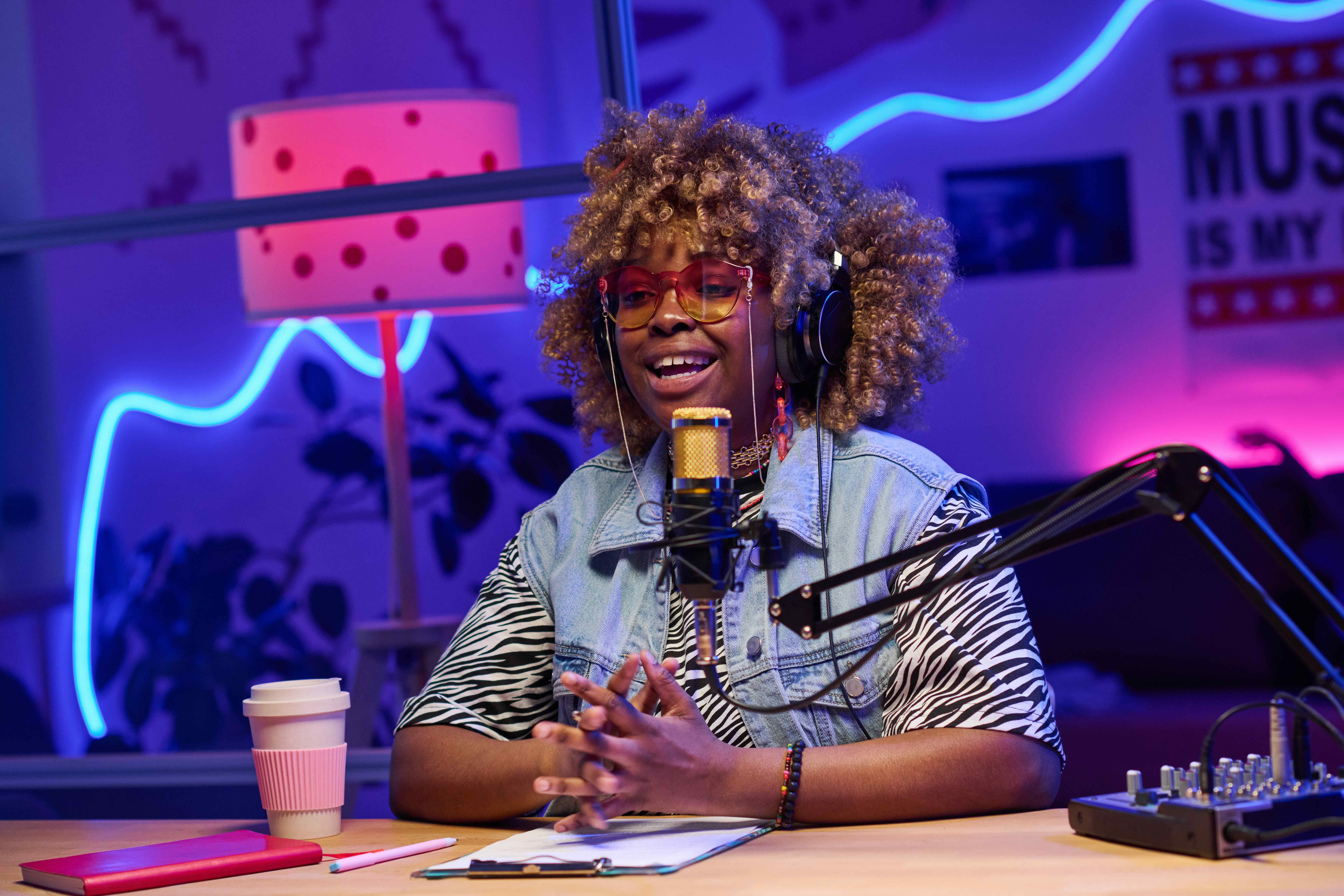 A young woman with voluminous blonde curls and tinted glasses sits at a desk in a vibrant, neon-lit studio, speaking into a professional microphone. She is wearing a denim vest over a zebra-print shirt and large headphones, appearing to host a podcast or r