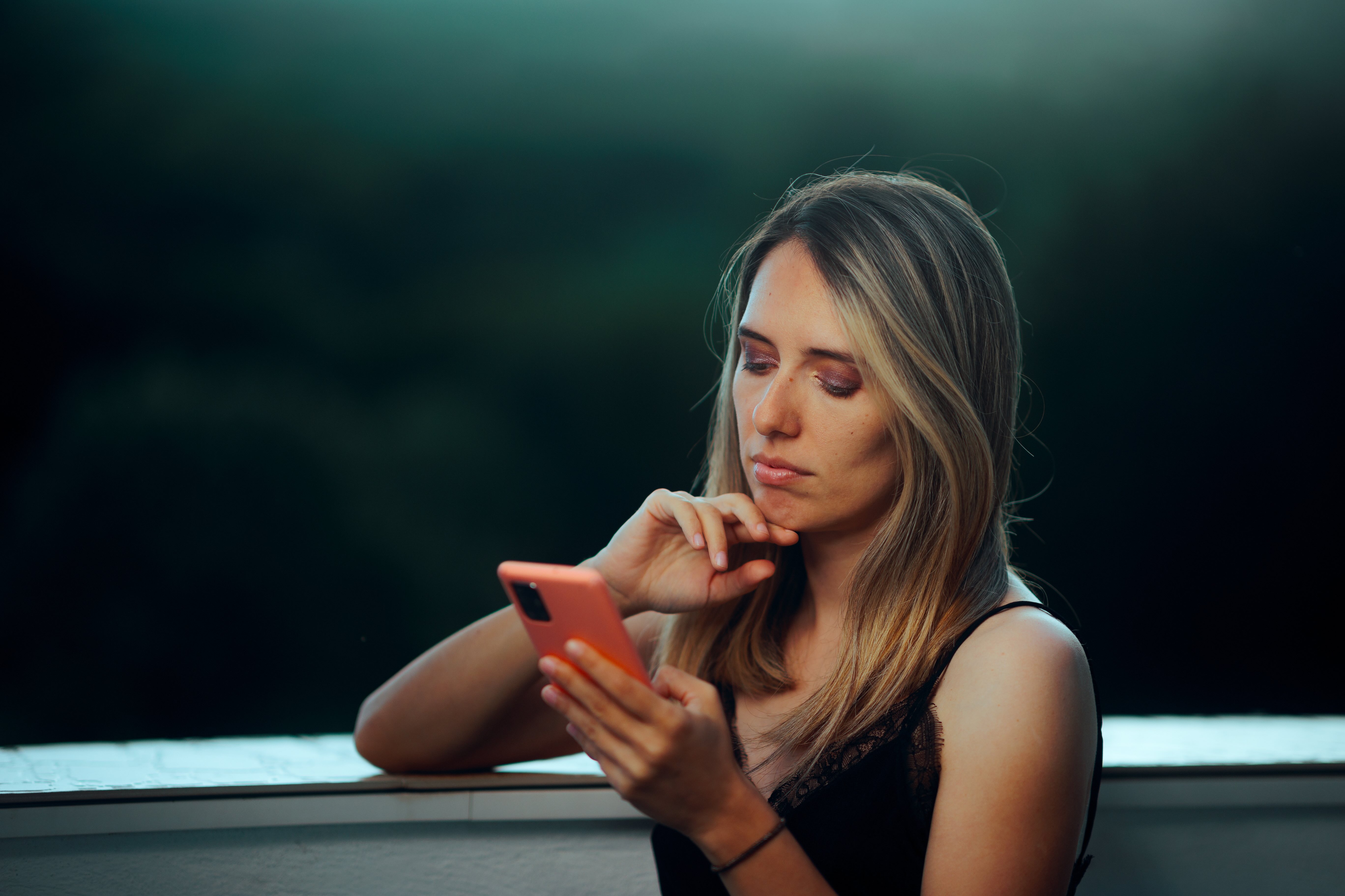  Gemini said In the image, a woman with long, blonde hair is looking down at a mobile phone with a contemplative and slightly somber expression. She is outdoors at dusk or in a low-light setting, leaning against what appears to be a balcony railing.