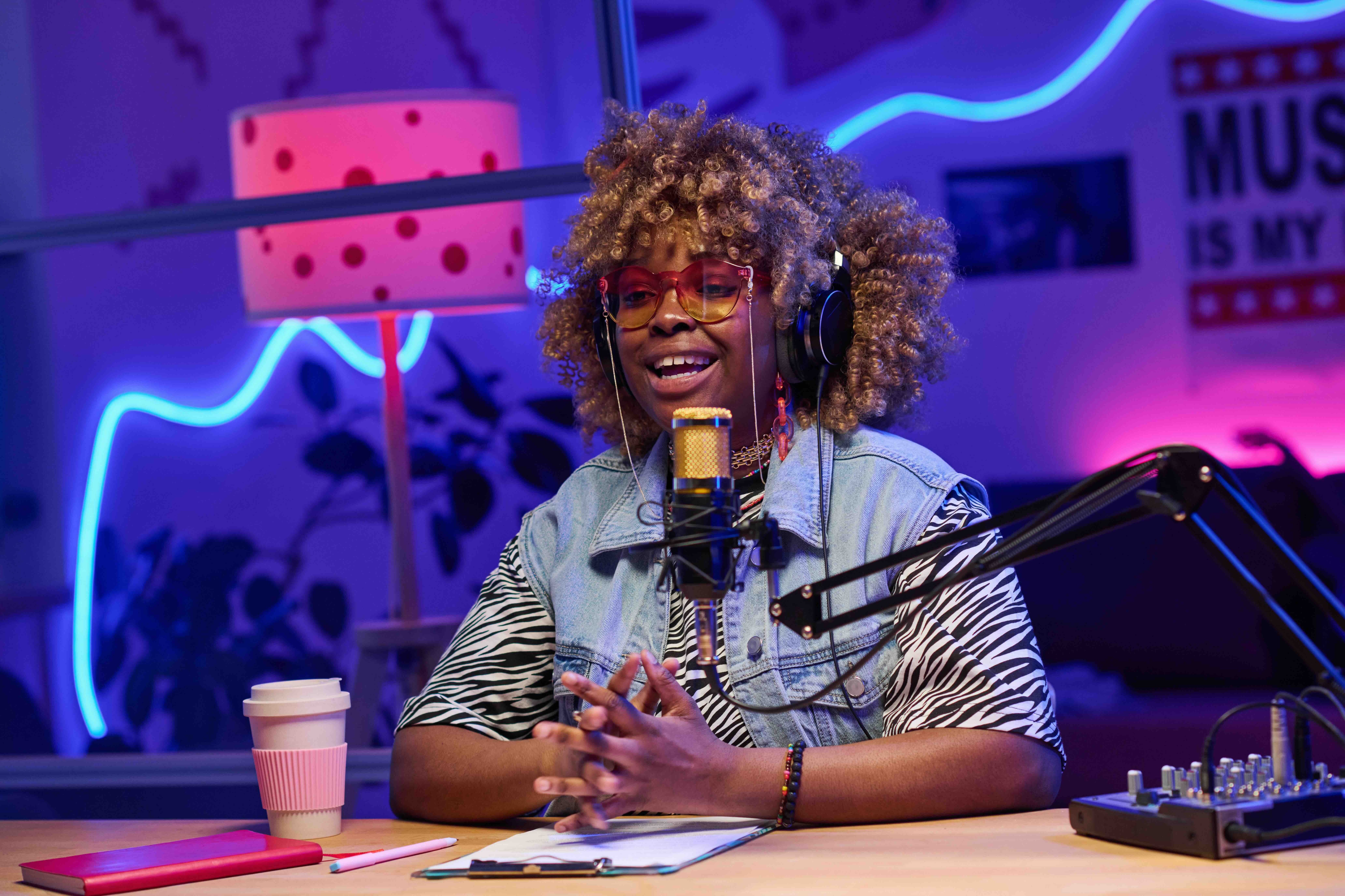 A medium-wide shot captures a female Black podcast host with blonde curly hair speaking with a smile, wearing oversized sunglasses with reddish-yellow tint and headphones while gesturing with her hands above notes on a clipboard and a microphone on a boom 
