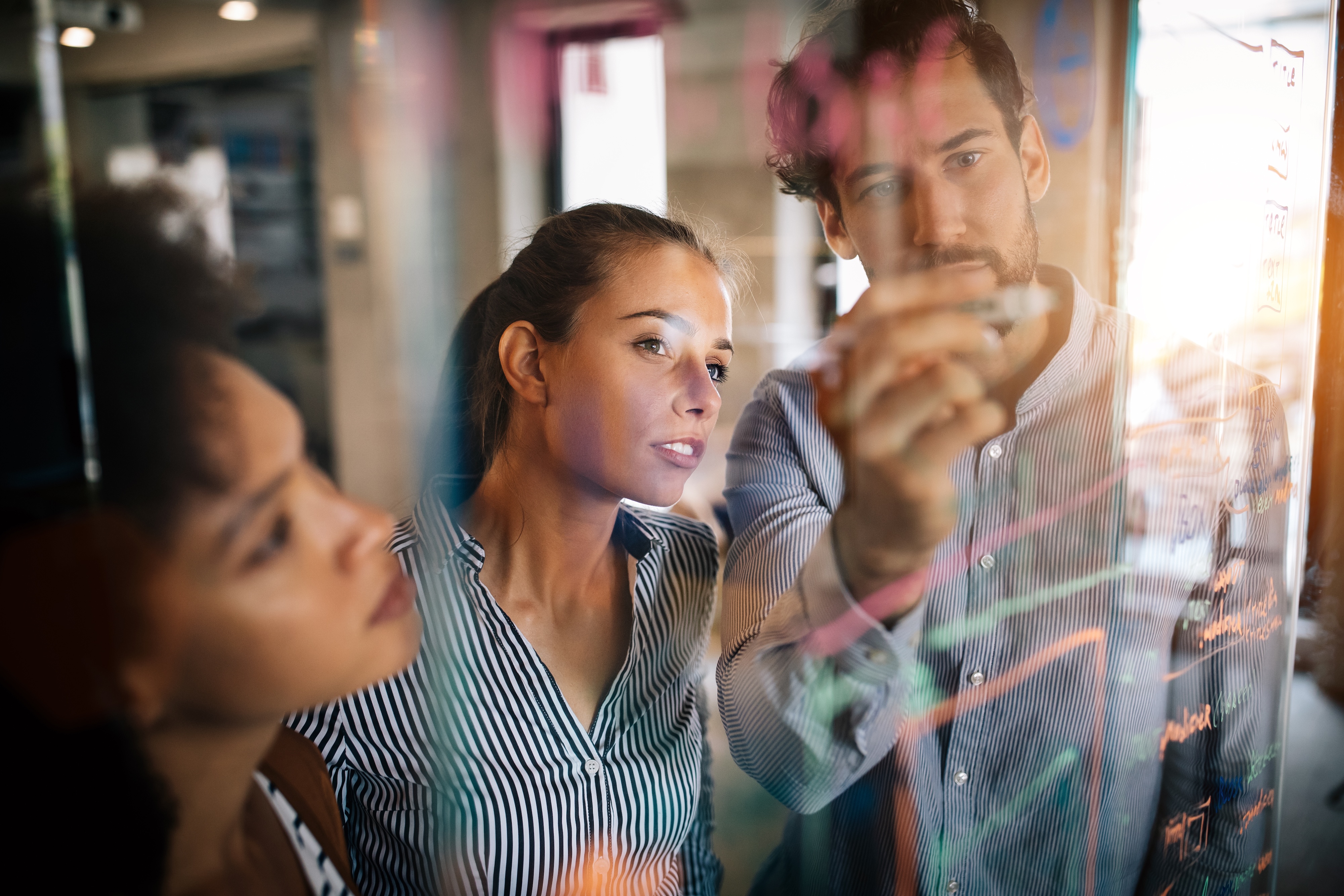A diverse team of three business professionals collaborates and discusses ideas while brainstorming on a clear glass whiteboard in a sunlit modern office. The man in the background writes with a marker as two women watch and listen attentively.