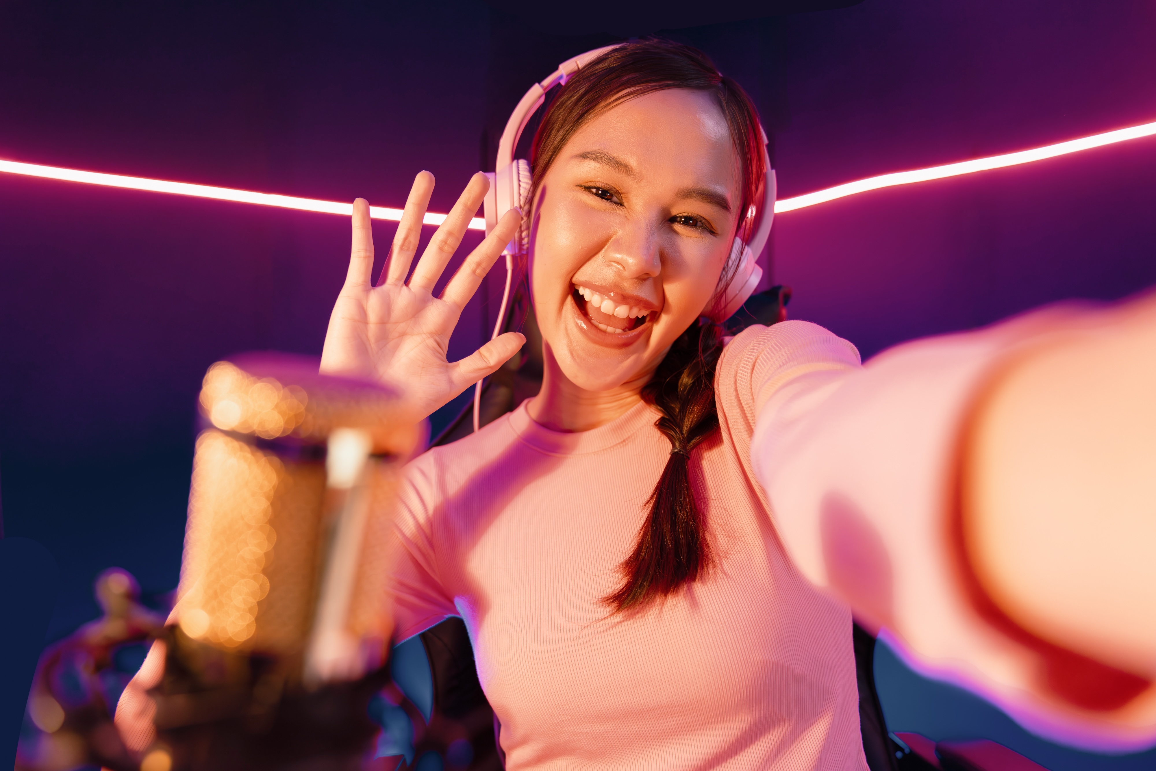A cheerful young Asian woman wearing white headphones, waving at the camera while taking a selfie in a neon-lit gaming room with a professional microphone in the foreground.