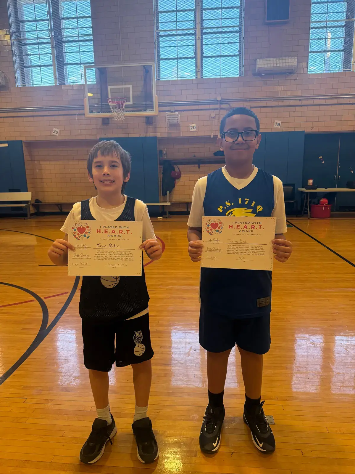 Two students holding I Played With HEART Award certificates in gymnasium
