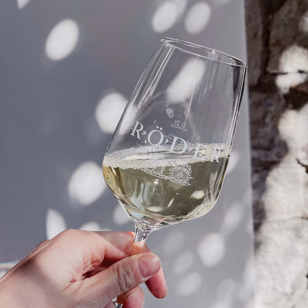Hand holding a tilted wine glass filled with white wine against a background with soft light and shadow patterns.