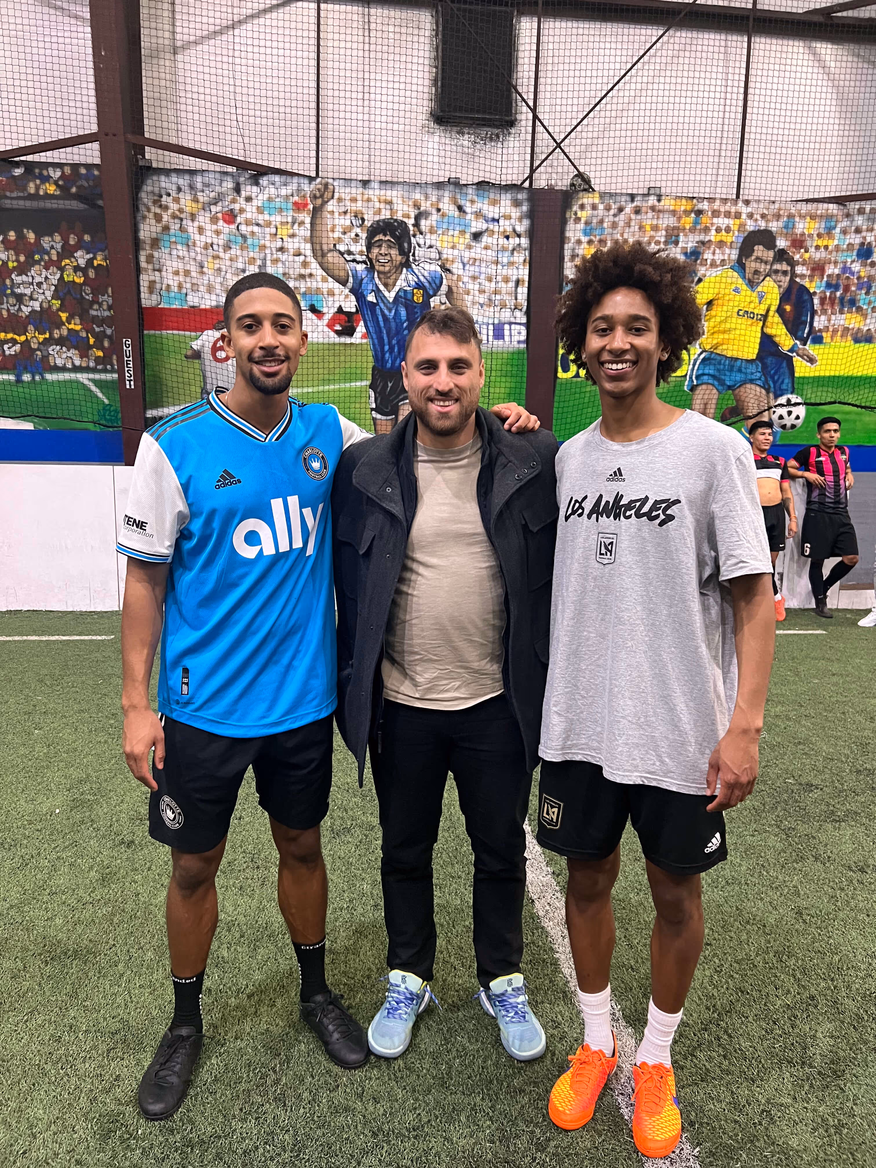 Three men standing on an indoor soccer field with painted murals of soccer players on the wall behind them.