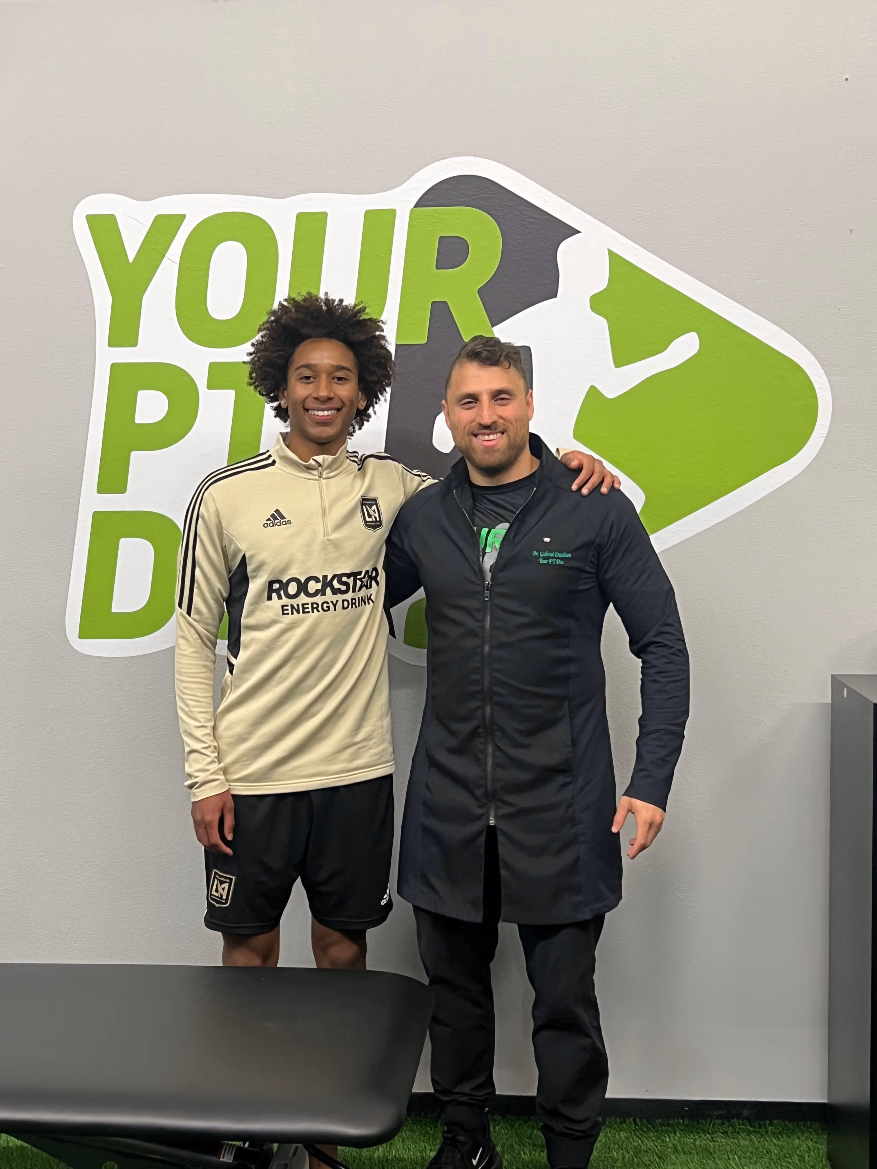 Two men smiling and posing together in front of a wall with green and black text reading 'YOUR PT DOC'. One wears an LAFC soccer jersey and shorts, the other wears a black coat with 'Dr. Gabriel Davidson Your PT Doc' embroidered on it.