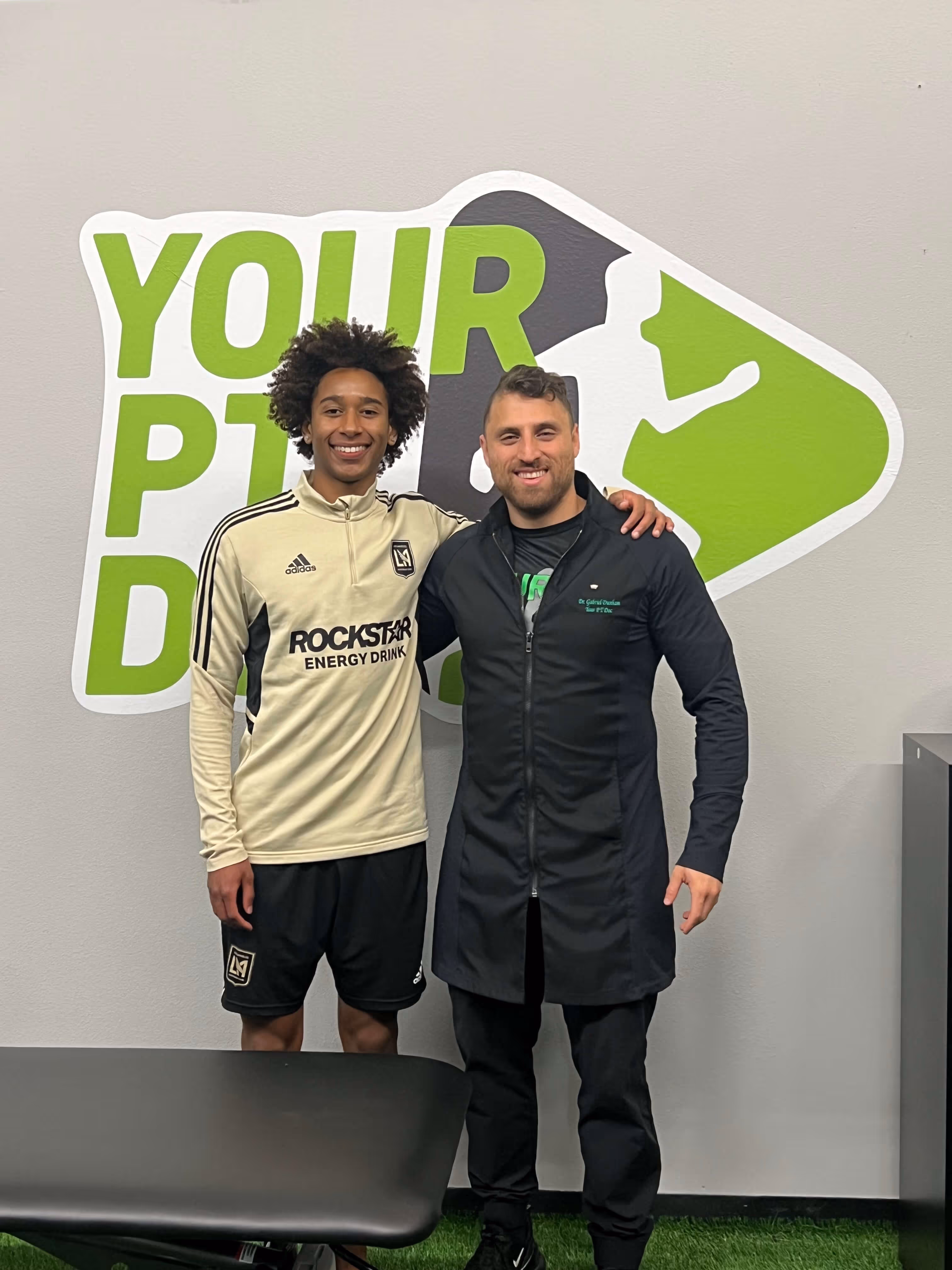 Two men smiling and posing with arms around each other in front of a wall with a large green and white fitness logo.